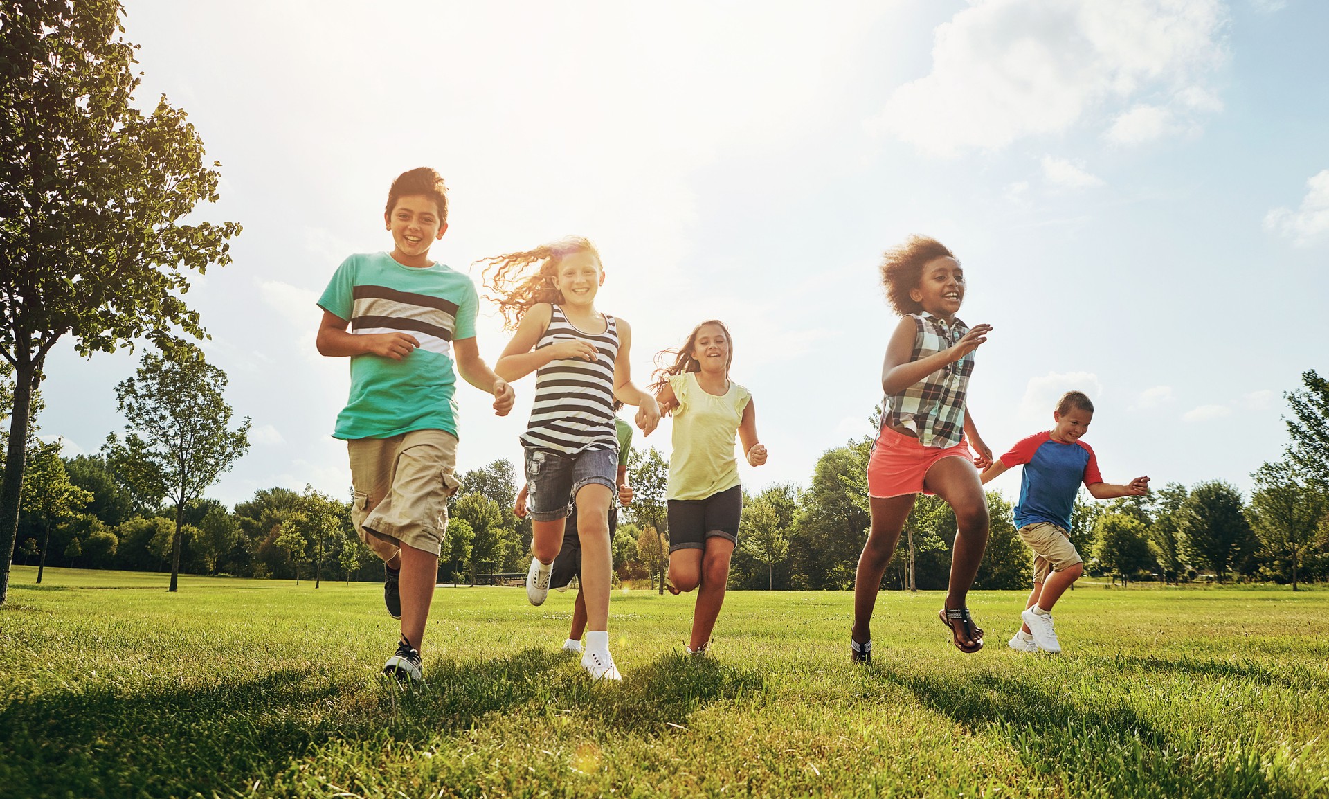 Happy friends, children and running with grass field in nature for fun, playful day or sunshine at park. Diversity or group of excited kids or youth enjoying sunny outdoor holiday or weekend together